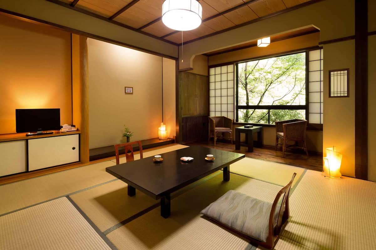 A traditional Japanese room with tatami flooring, a low table with cushions, a television on a cabinet, and a view of trees through shoji screens.
