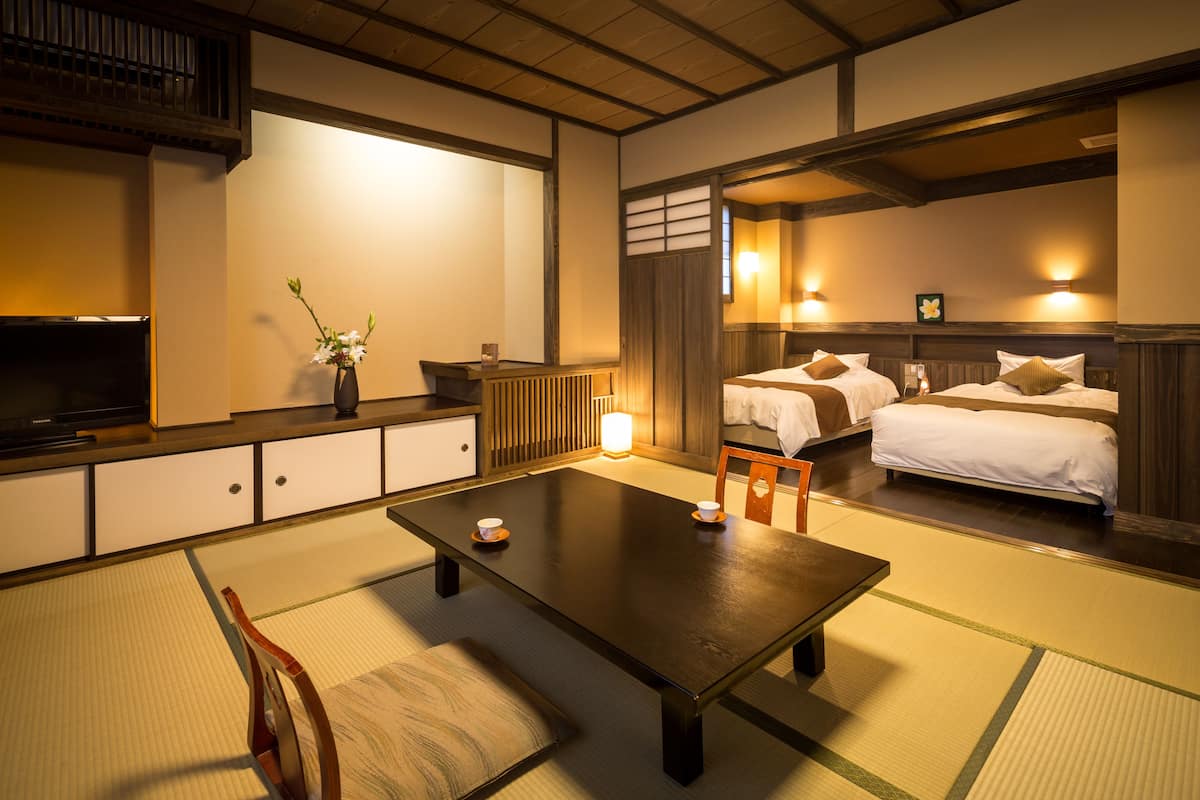 A traditional Japanese-style room with tatami flooring, a low table, a TV, and two beds with pillows and a nightstand.