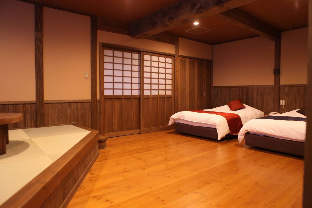 A traditional Japanese-style room with tatami flooring, two beds with white bedding and red accents, a wooden table, and sliding shoji doors.