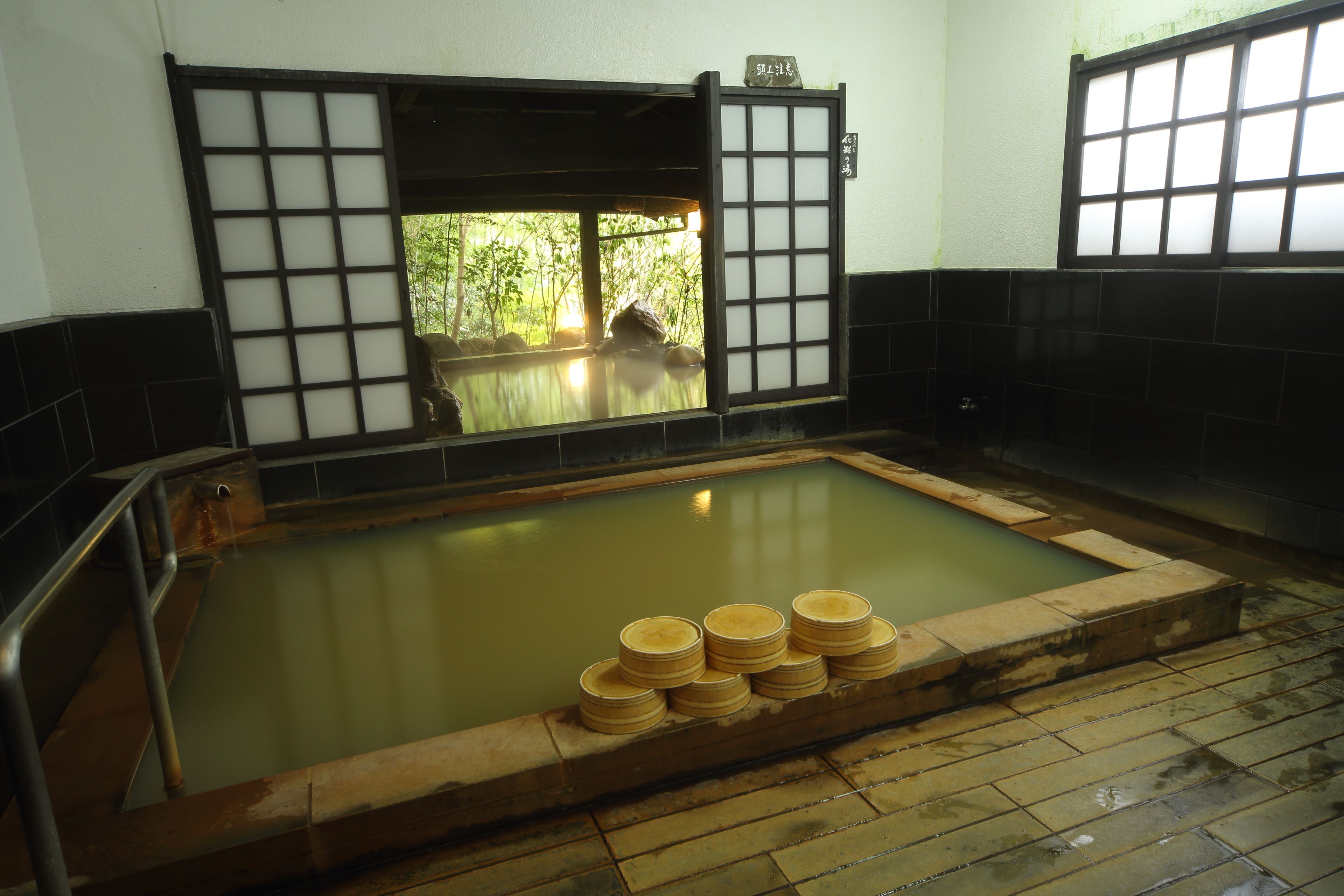 A traditional Japanese onsen with a wooden bath surrounded by tatami mats, a stone platform with wooden buckets, and a view of greenery through a shoji screen.