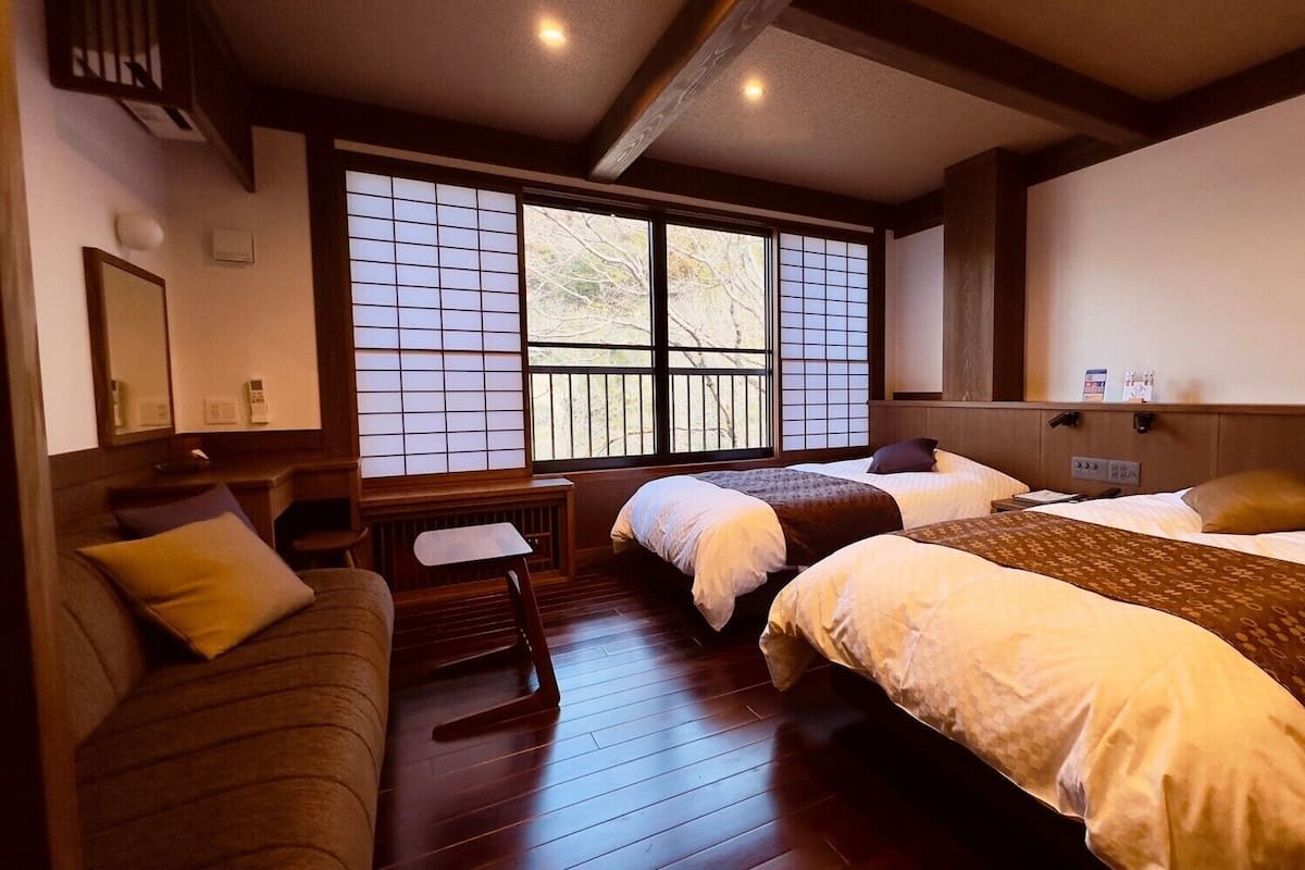 A hotel room with two beds, a small table, a sofa, and a large window with a view.