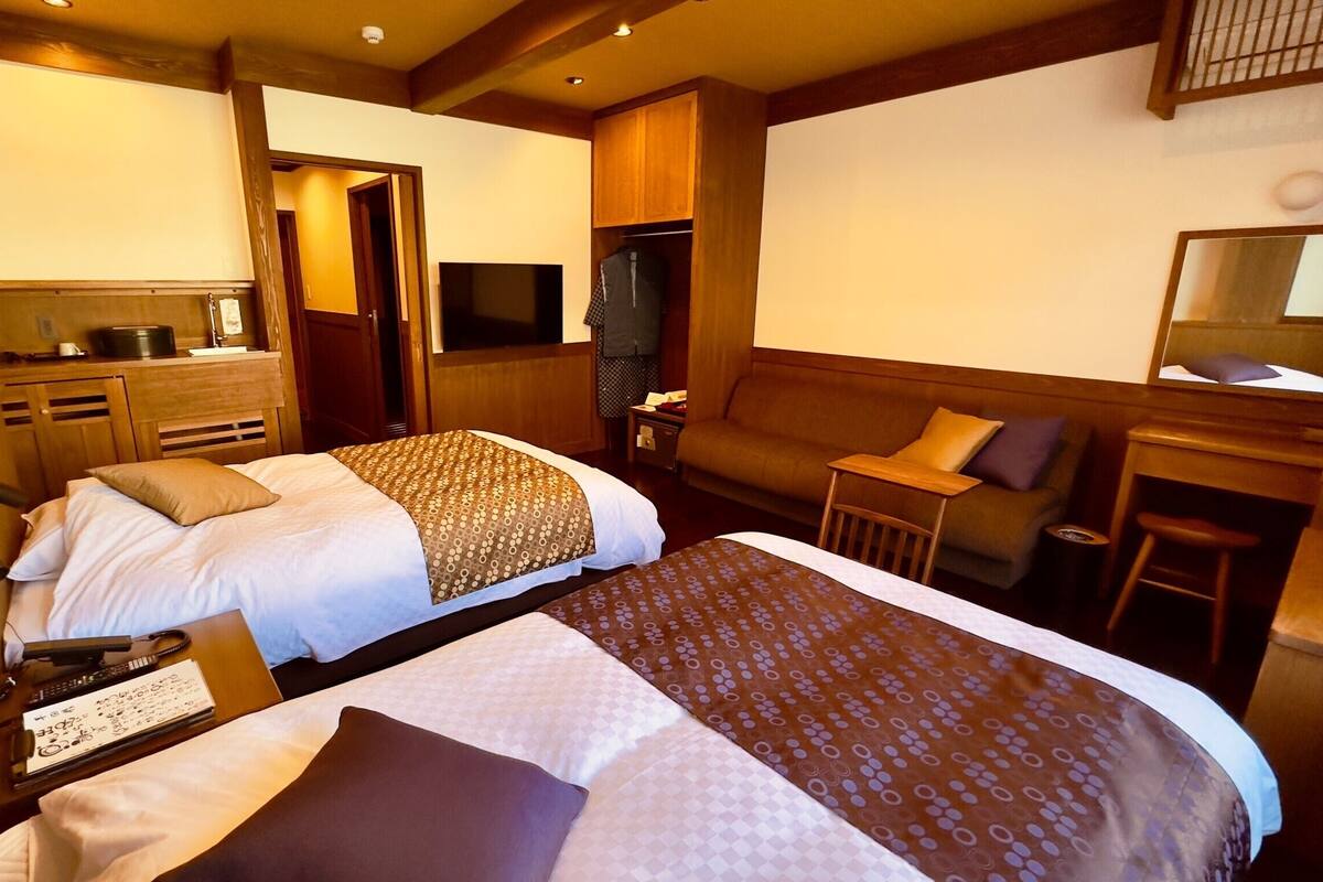 A hotel room with two beds, a desk, a chair, a sofa, and a television.