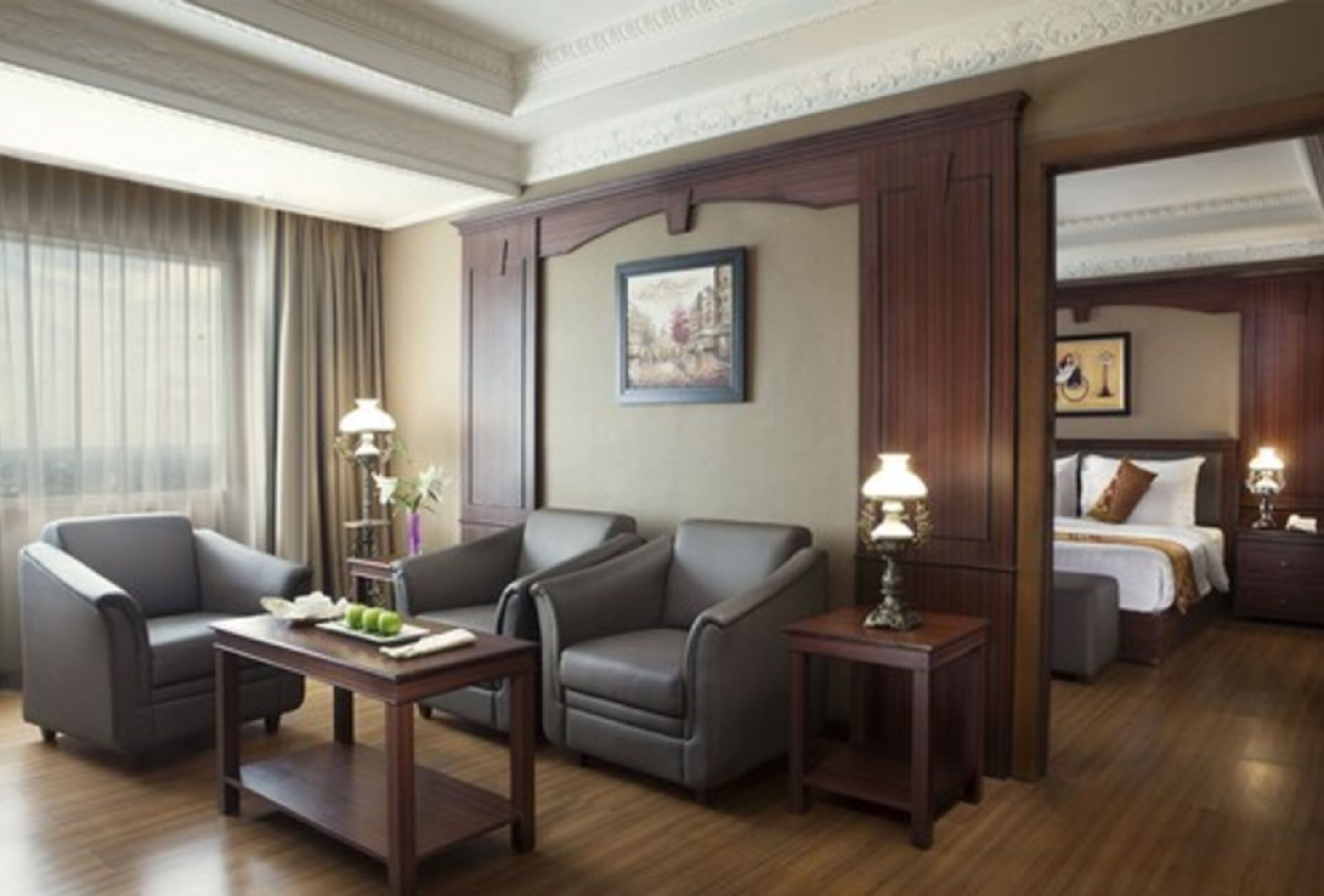 A hotel room with a large bed, two armchairs, a small table, and a desk. There is a painting on the wall and a window with curtains.