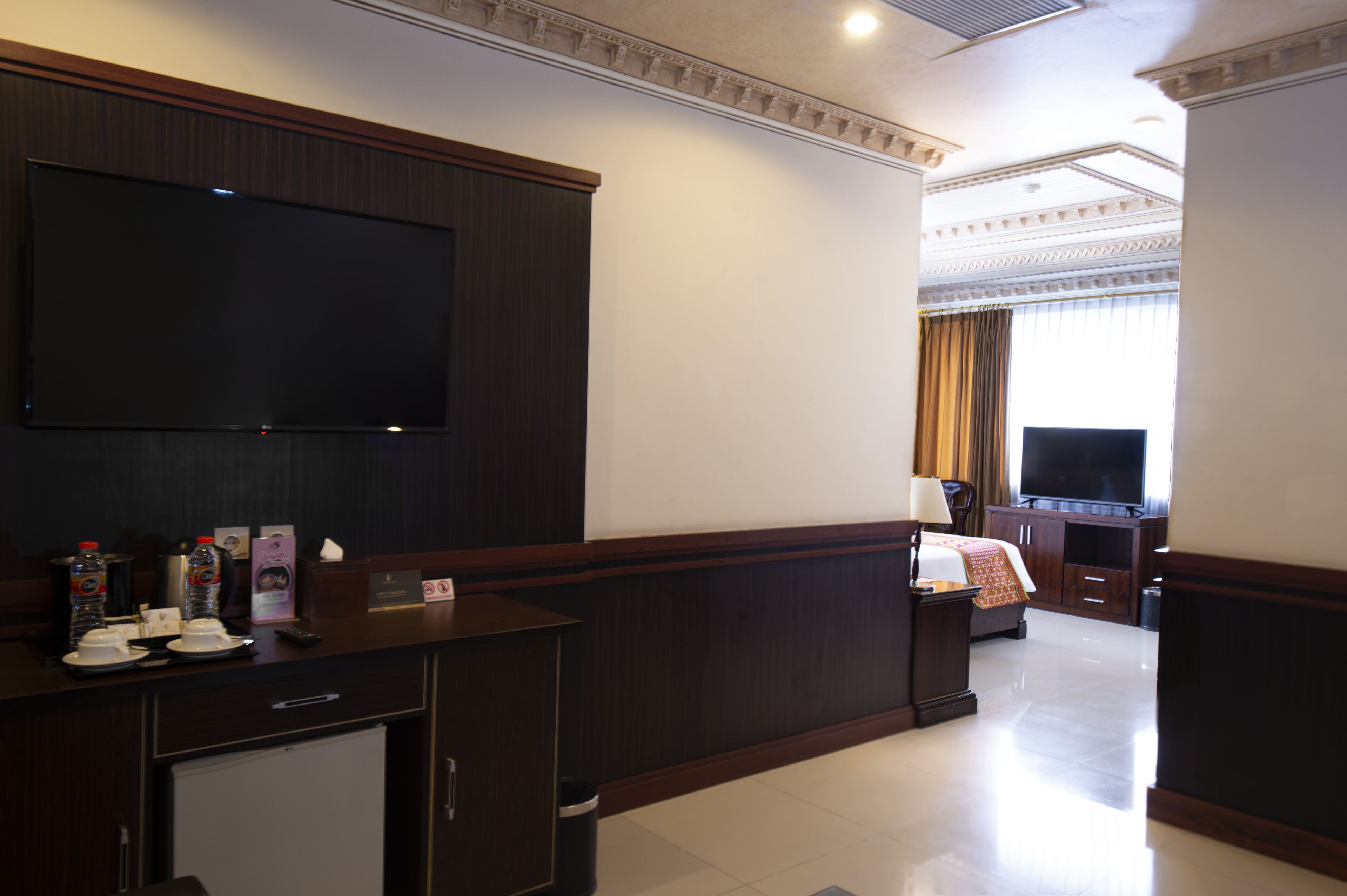 A hotel room with a flat-screen TV, a desk with a lamp, a bed, and a large window with curtains.