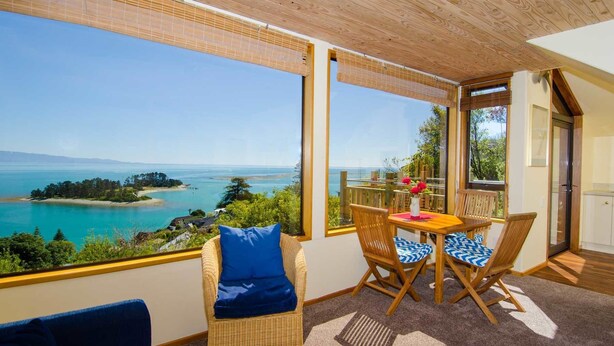 A room with a view of a coastal landscape, featuring a table with chairs, a wicker sofa, and a wooden ceiling. - The Wheelhouse Inn (Nelson)