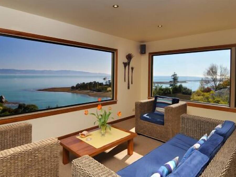 A modern living room with a large window overlooking a scenic coastal view. There is a wooden coffee table, wicker furniture, and a blue sofa.
