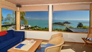 A room with a blue sofa, a wooden coffee table, and a wicker chair. Large windows offer a view of the sea and a small island. - The Wheelhouse Inn (Nelson)