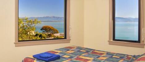 A bedroom with two windows offering a view of a lake and mountains.