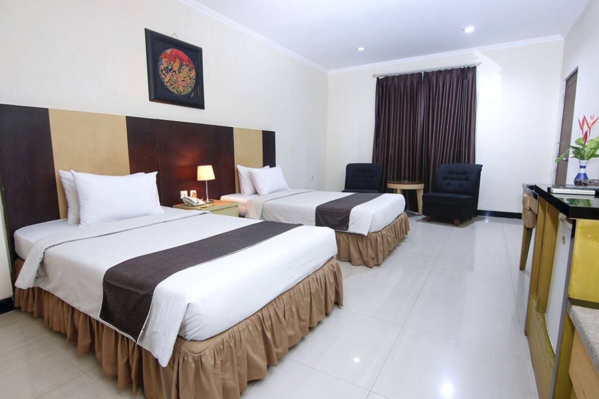 A hotel room with two beds, a desk, a chair, a lamp, and a painting on the wall.