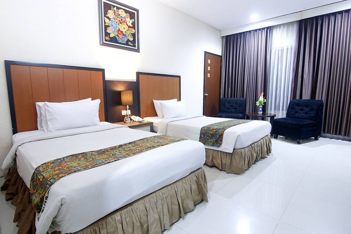 A hotel room with two beds, a large window with curtains, a chair, and a painting on the wall.