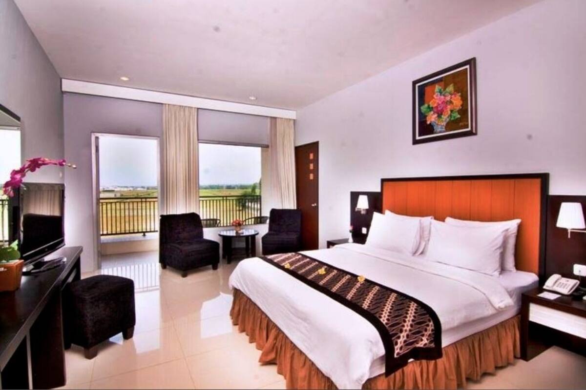 A hotel room with a large bed, a desk with a computer, a chair, and a balcony with a view of the countryside.