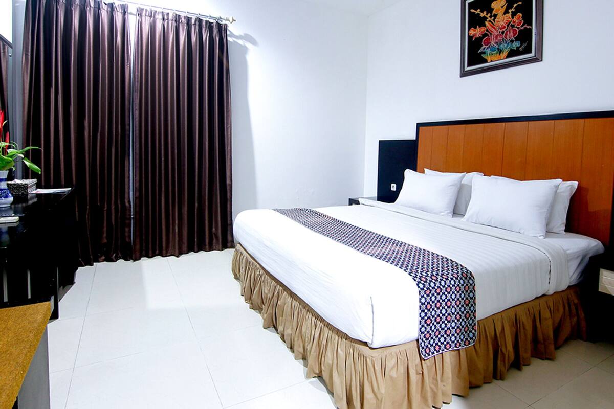 A hotel room with a large bed, a wooden headboard, a nightstand, a desk with a plant, and a window with curtains.