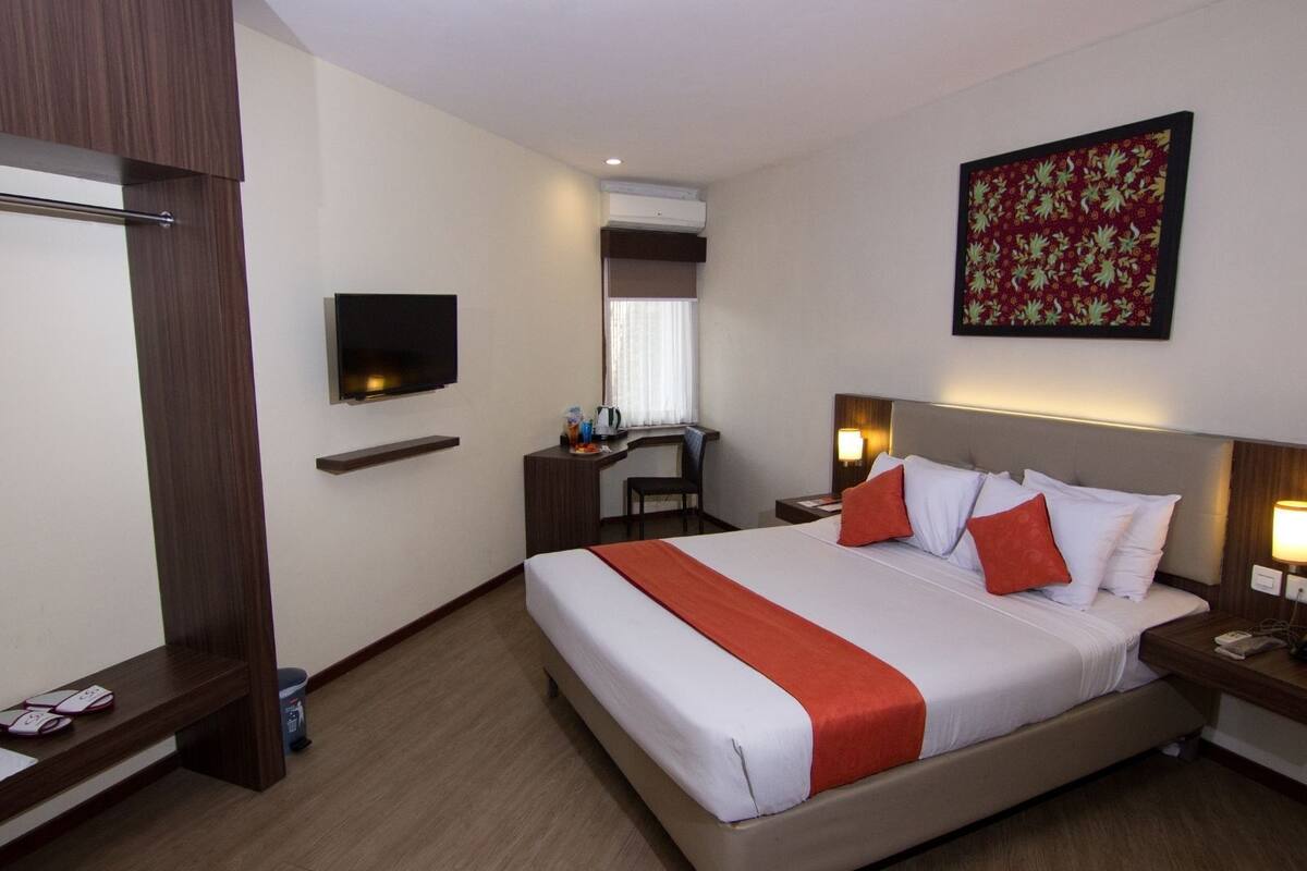 Quarto de hotel com cama, escrivaninha, televisão e um quadro na parede.