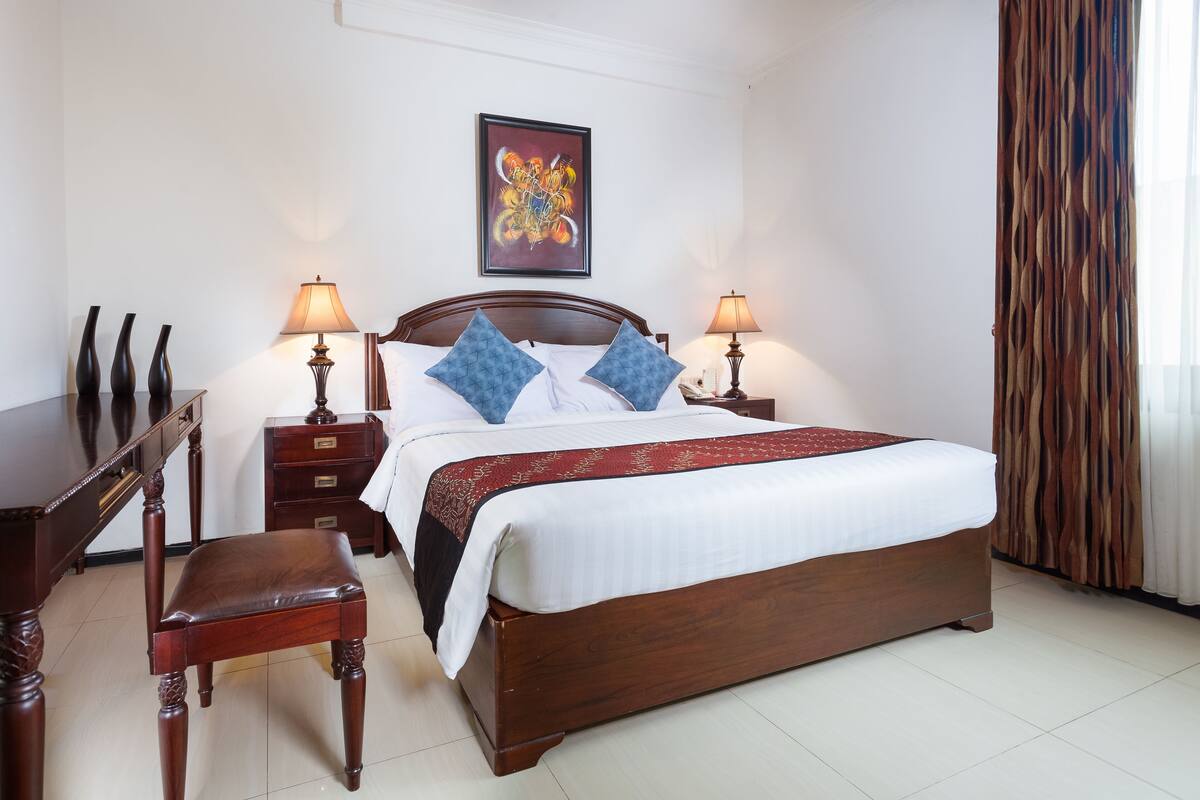 Quarto de hotel bem arrumado com uma cama de madeira, duas mesas de cabeceira com luminárias, uma escrivaninha e uma cadeira.