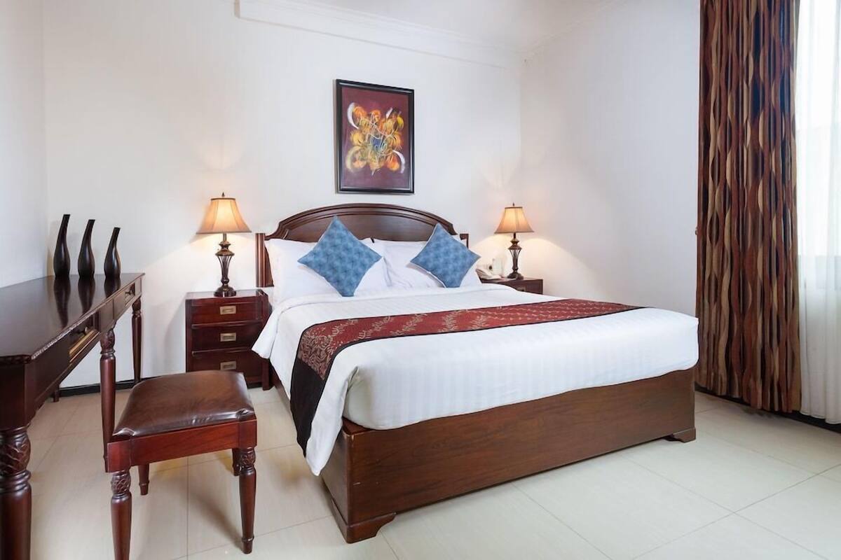 Quarto de hotel bem arrumado com uma cama de madeira, duas mesas de cabeceira com luminárias, uma escrivaninha e uma cadeira.