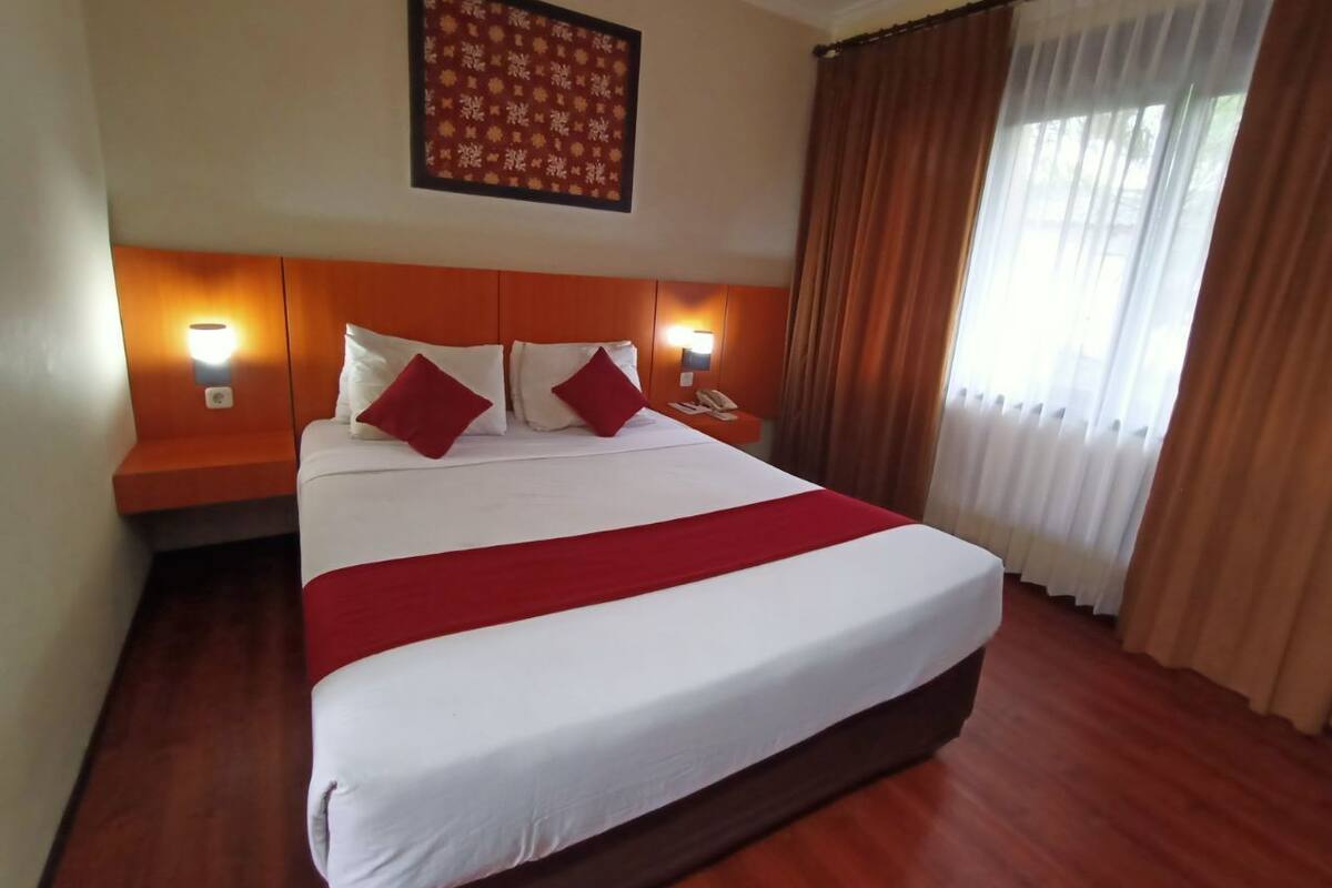 Quarto de hotel com uma cama grande, duas mesas de cabeceira com luminárias, cabeceira, janela com cortinas e um enfeite na parede.