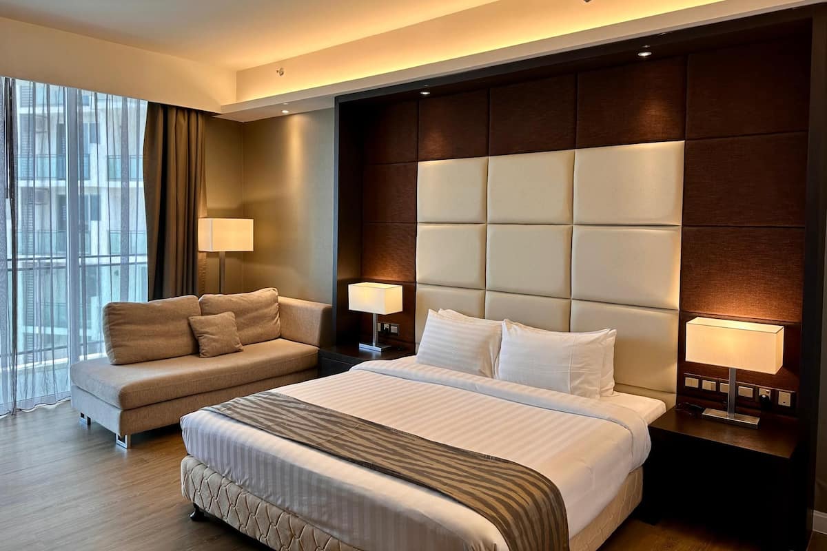Una habitación de hotel moderna con una cama grande, un sofá, dos lámparas de noche y vistas a la ciudad a través de amplios ventanales.