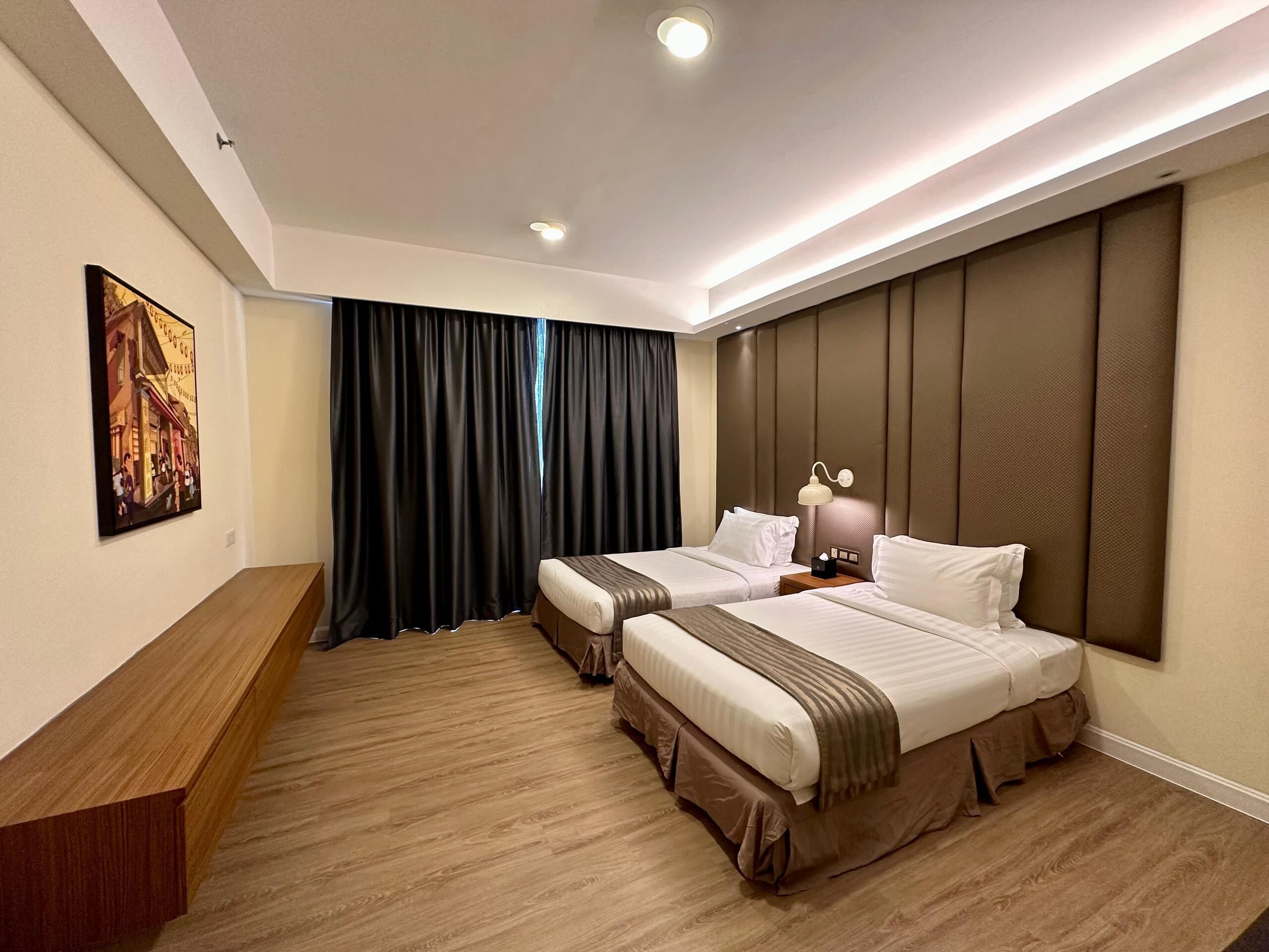 A hotel room with two beds, a wooden desk, a painting on the wall, and a large window with curtains.