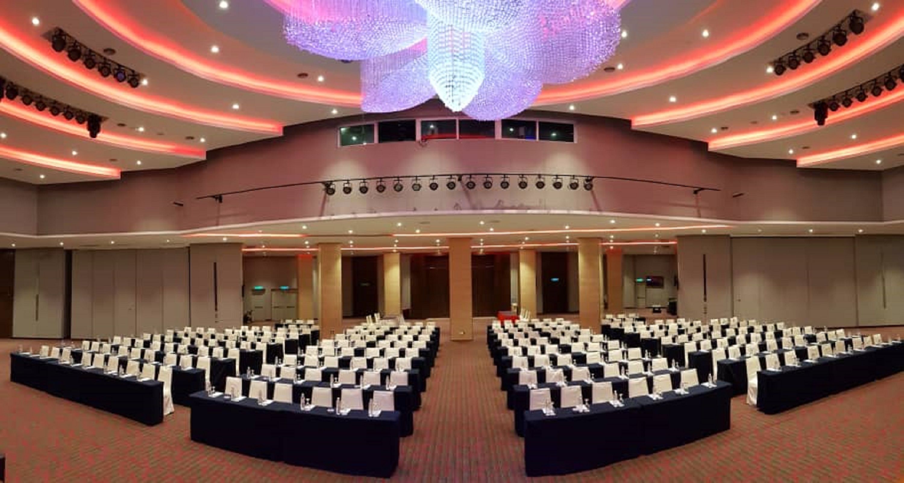 Un amplio salón de conferencias con filas de mesas y sillas, un techo alto con una gran lámpara decorativa y un escenario con equipo de iluminación.