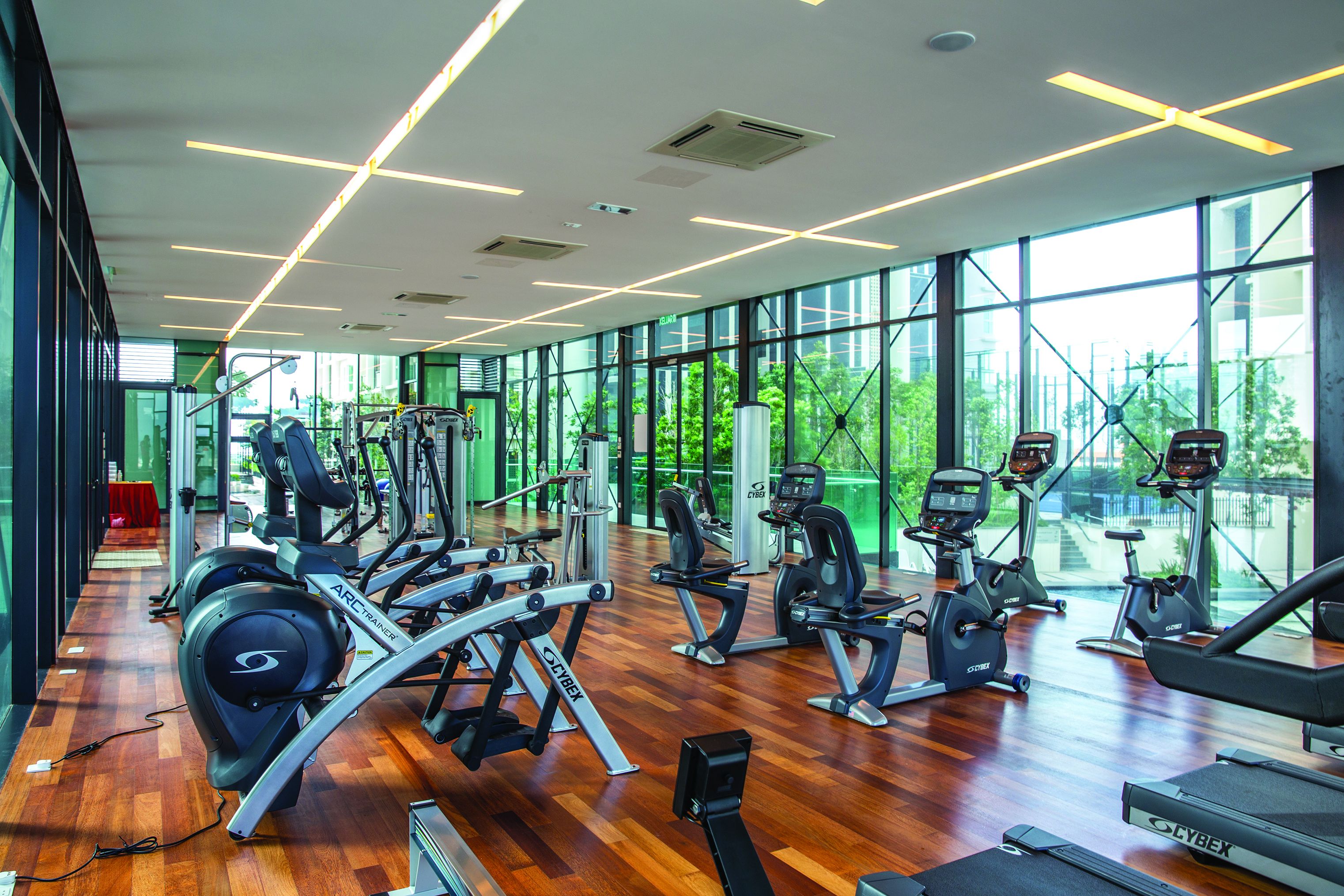 A modern gym with cardio machines, weight stations, and large windows offering a view of greenery.