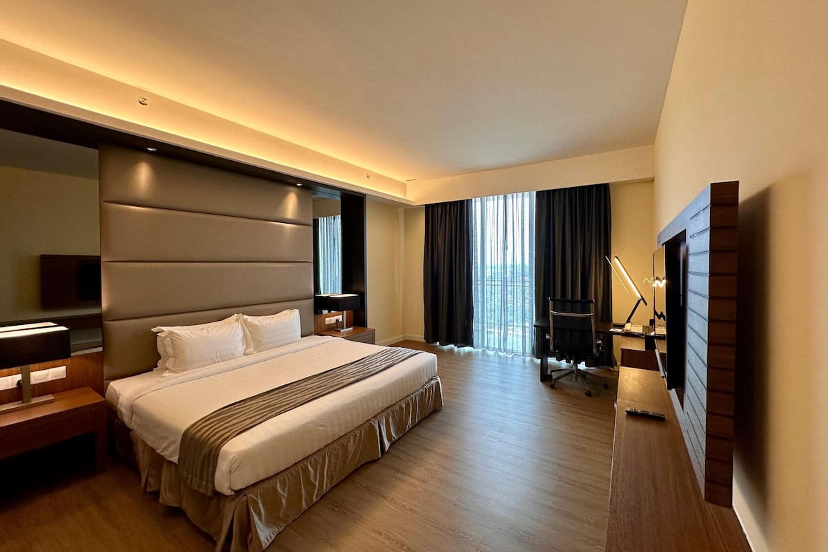 Una habitación de hotel moderna con una cama grande, un escritorio, una silla y un amplio ventanal con cortinas.