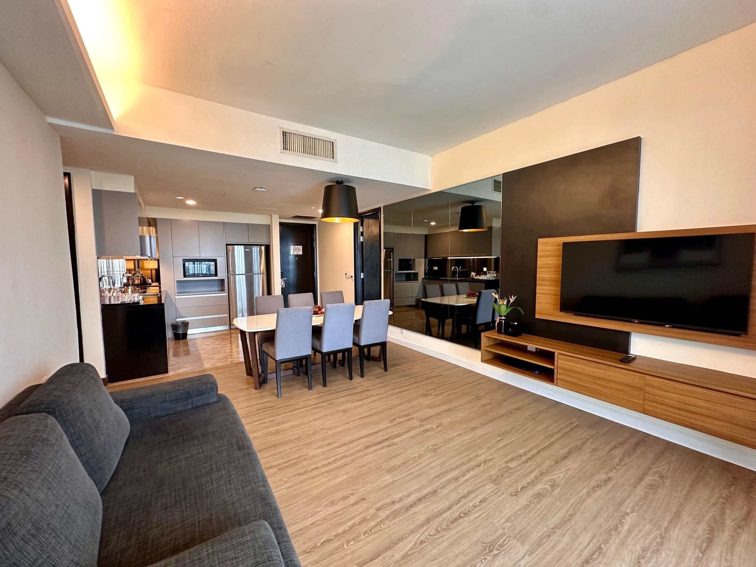 A modern living room with a grey sofa, a dining area with a table and chairs, a kitchen in the background, and a large flat-screen TV mounted on the wall.