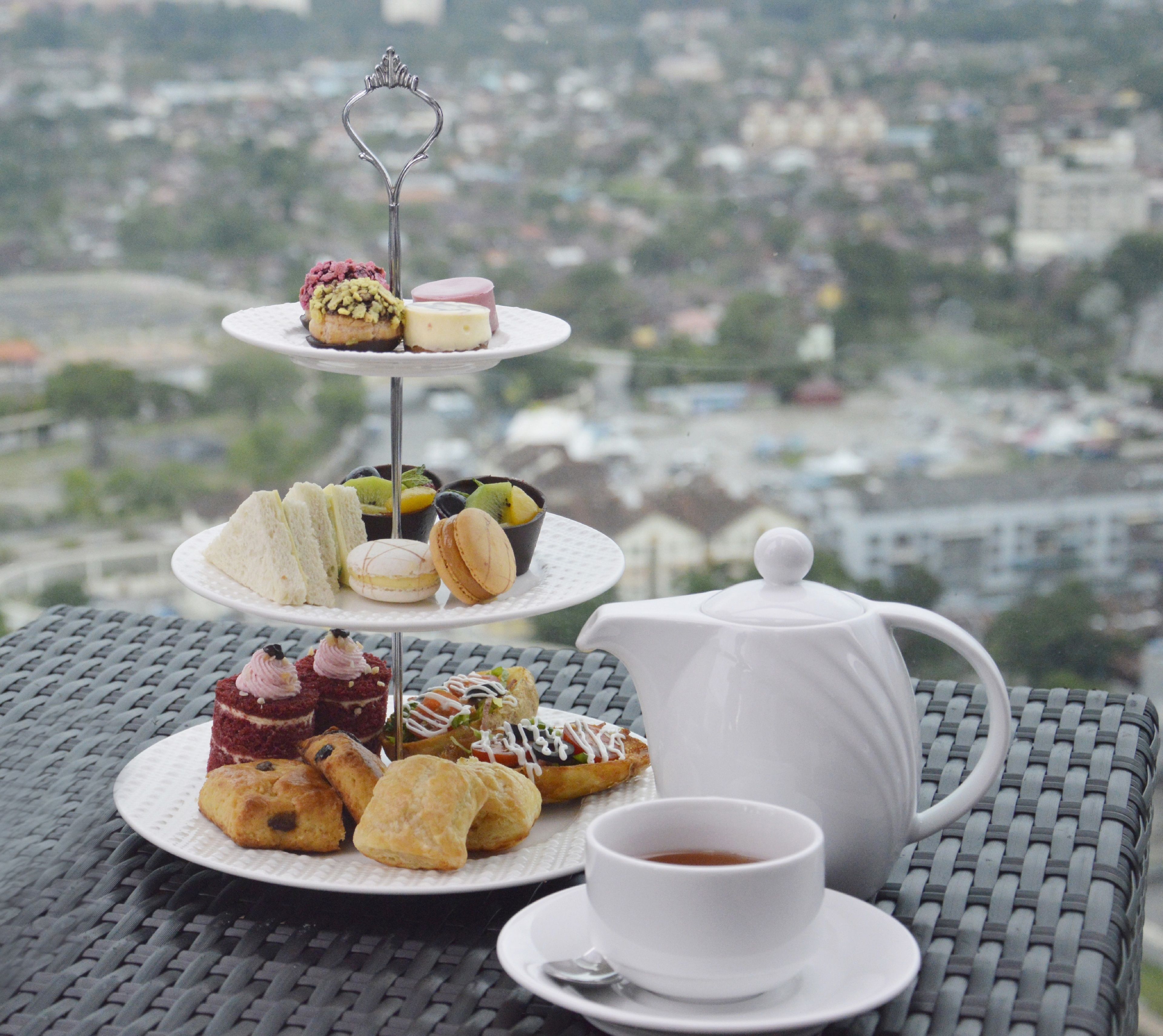 Un expositor escalonado para postres con varios pasteles y una tetera sobre una mesa, con vistas a la ciudad de fondo.