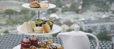 Un expositor escalonado para postres con varios pasteles y una tetera sobre una mesa, con vistas a la ciudad de fondo.