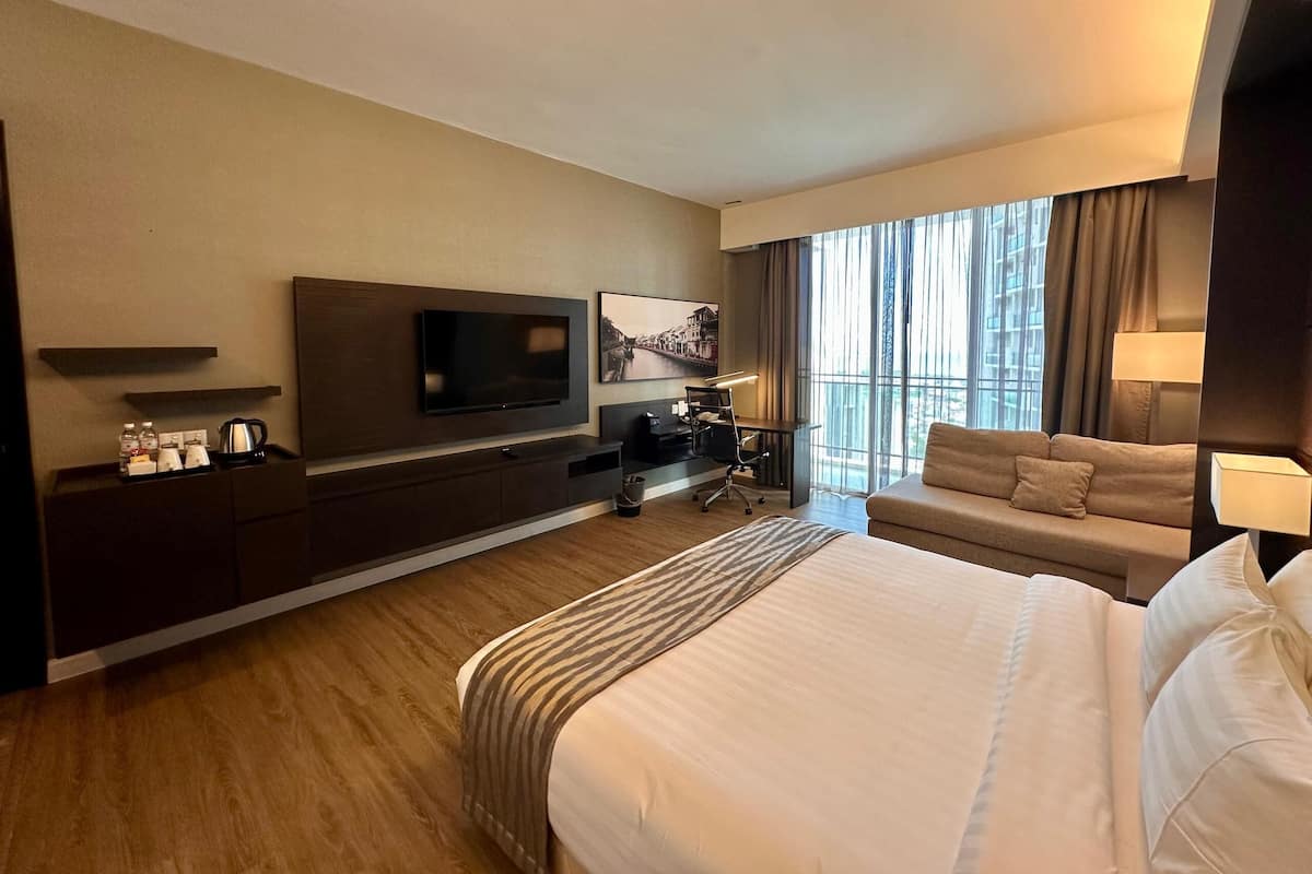 Habitación de hotel con una cama grande, un televisor montado en la pared, un escritorio con una silla, un sofá y una ventana con cortinas.