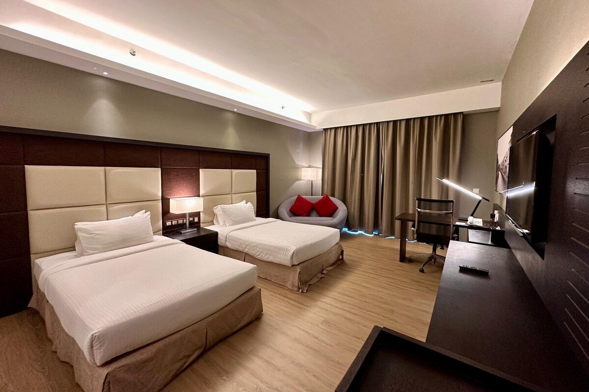 Ein Hotelzimmer mit zwei Betten, einem Schreibtisch, einem Stuhl, einem Fernseher und einem großen Fenster mit Vorhängen.