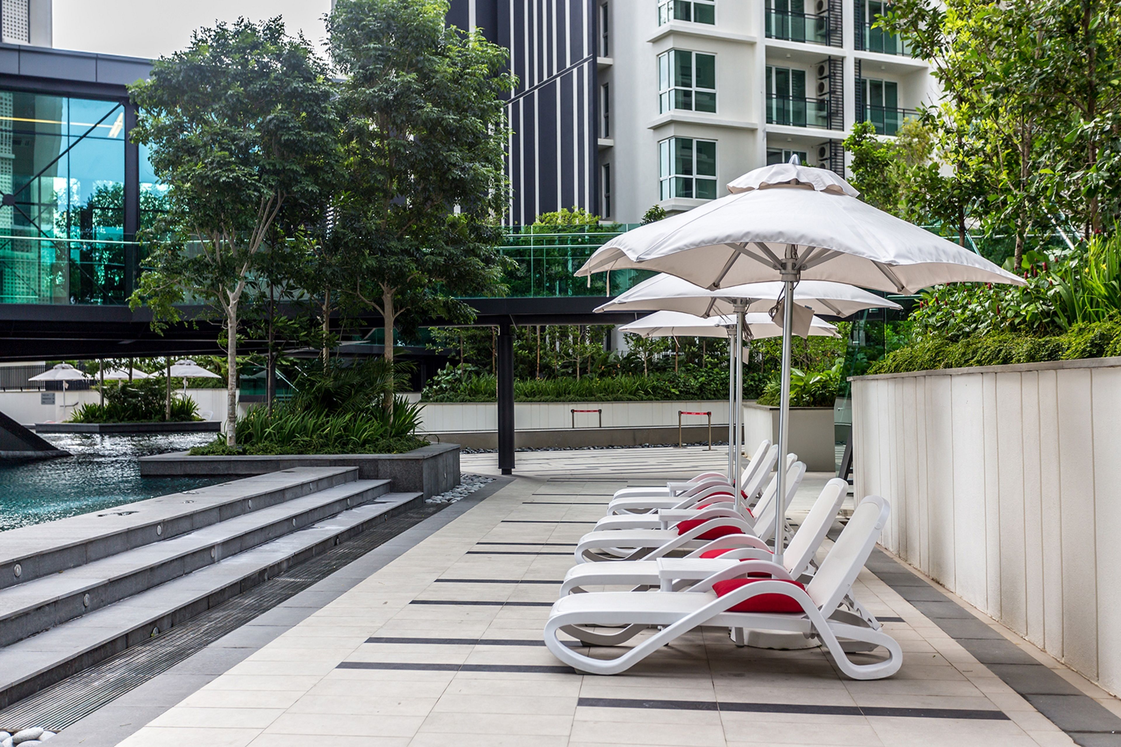 Un espace aménagé au bord de la piscine, avec des chaises longues et des parasols, une fontaine ou un autre élément décoratif aquatique, et un bâtiment moderne en arrière-plan.
