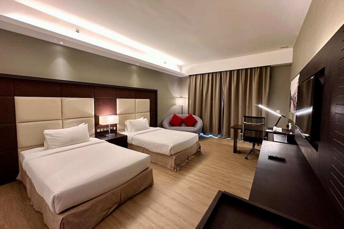 Habitación de hotel con dos camas, un escritorio, una silla, un televisor y un ventanal con cortinas.