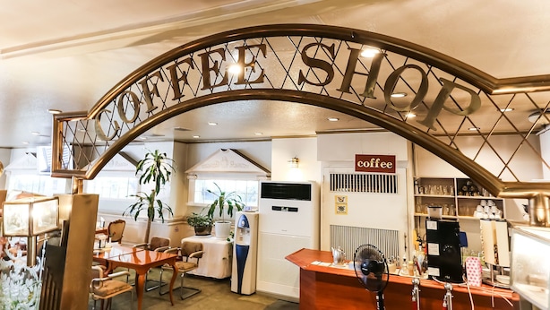Interior de una cafetería con un letrero que dice "COFFEE SHOP", una recepción, zona de estar y una máquina de café.