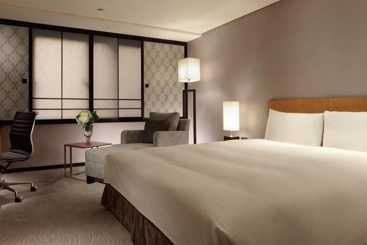 A hotel room with a large bed, a desk with a chair, a lamp, and a view of the city.
