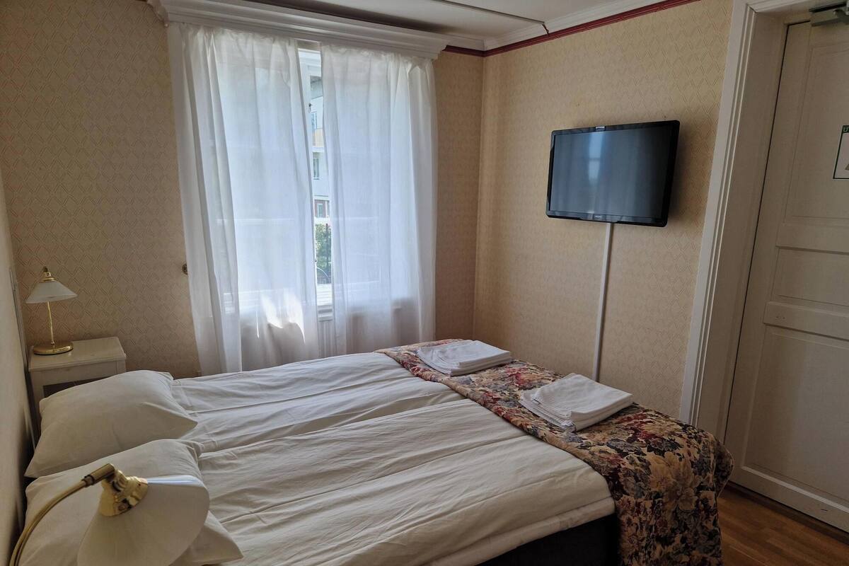 A hotel room with a bed, a TV mounted on the wall, a bedside lamp, and a window with sheer curtains.