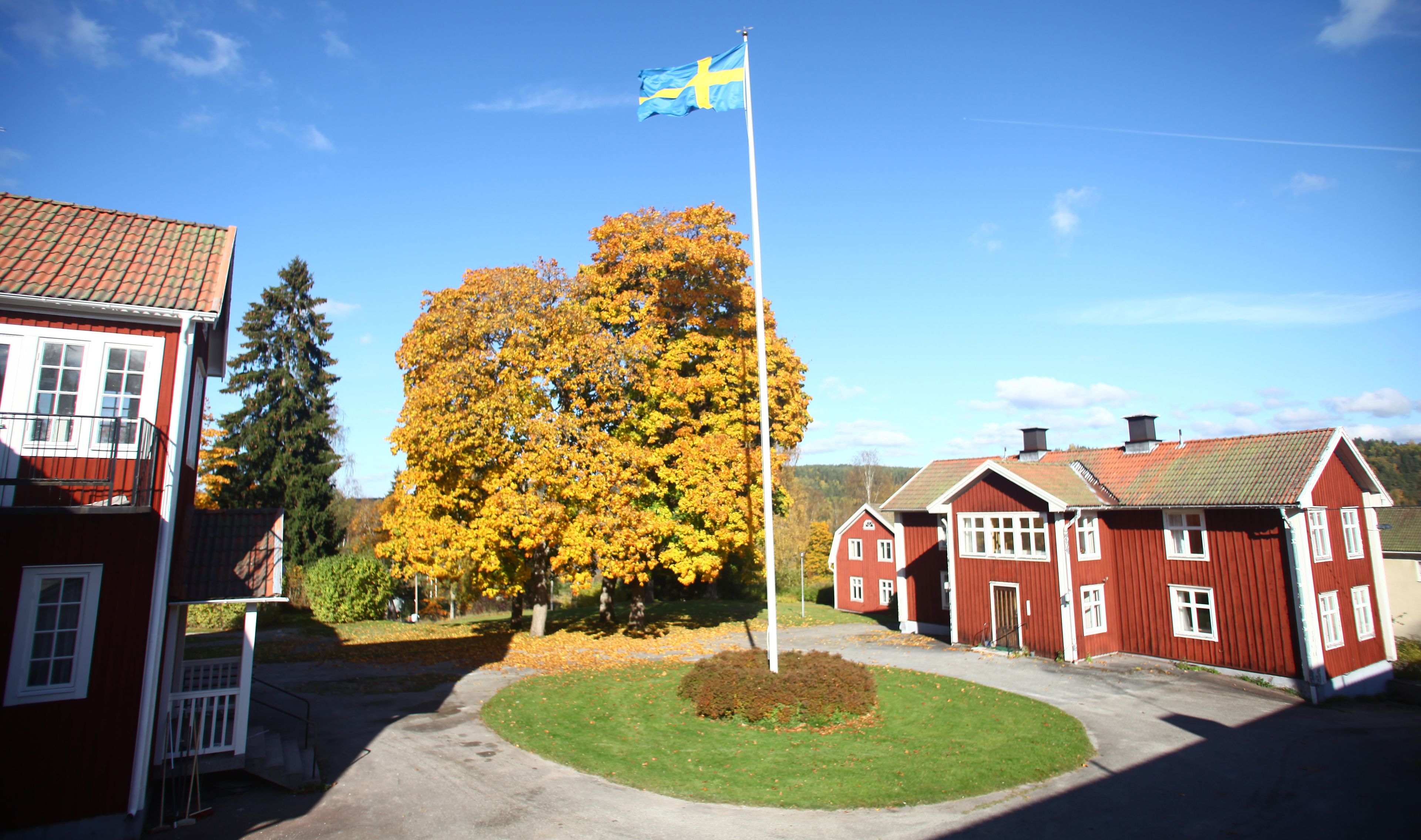 Un bâtiment rouge surmonté d’un mât portant un drapeau, un grand arbre aux feuilles jaunes et un ciel bleu dégagé.
