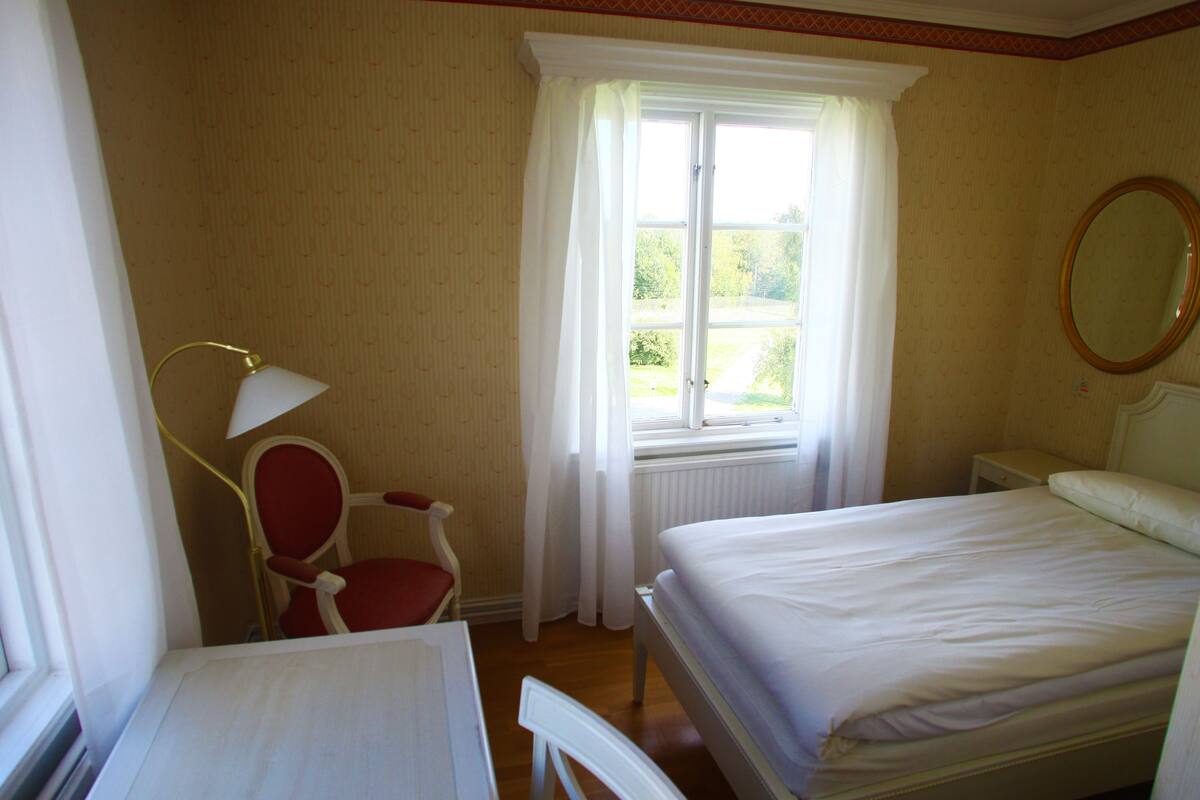 A bedroom with a bed, a chair, a desk, a window with curtains, and a mirror.
