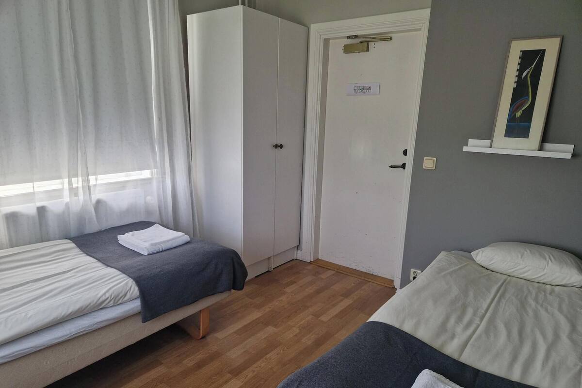 A room with two beds, a wardrobe, a door with a nameplate, and a wall-mounted shelf with a picture.