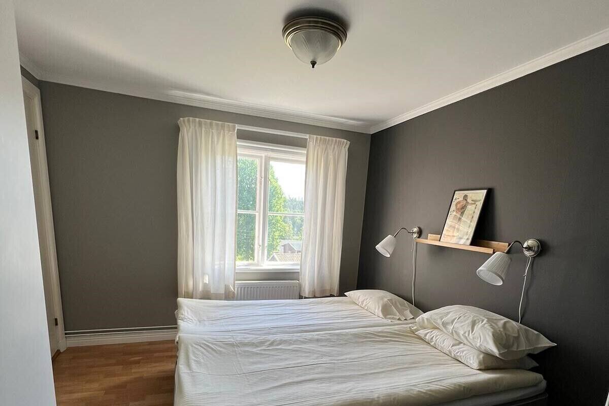 A bedroom with a bed, a window with curtains, a bedside table, and a wall-mounted lamp.