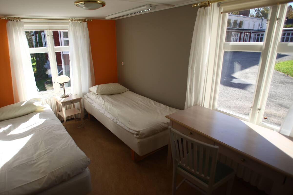 A small, well-lit room with two single beds, a nightstand with a lamp, and a window with curtains.