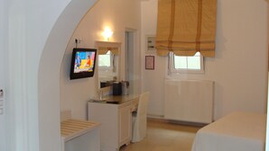 A hotel room with a white arched doorway, a desk with a chair, a bed, a window with a curtain, and a television on the wall. - Villa Christina (Skiathos)