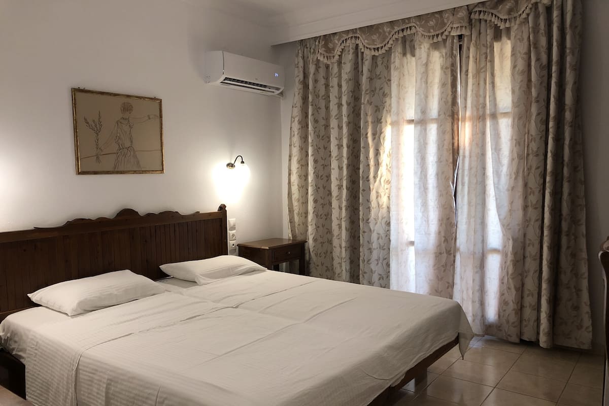 A neatly made bed with white linens, a wooden headboard, a wall-mounted air conditioner, a bedside table with a lamp, and a window with floral curtains.