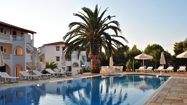 A swimming pool with lounge chairs and umbrellas, surrounded by buildings with balconies and palm trees. - Villa Christina (Skiathos)