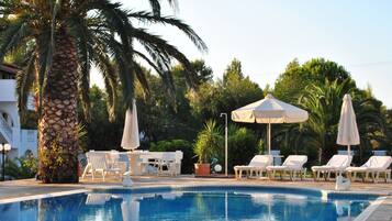 Una piscina con sillas de descanso y sombrillas, rodeada de palmeras y follaje.