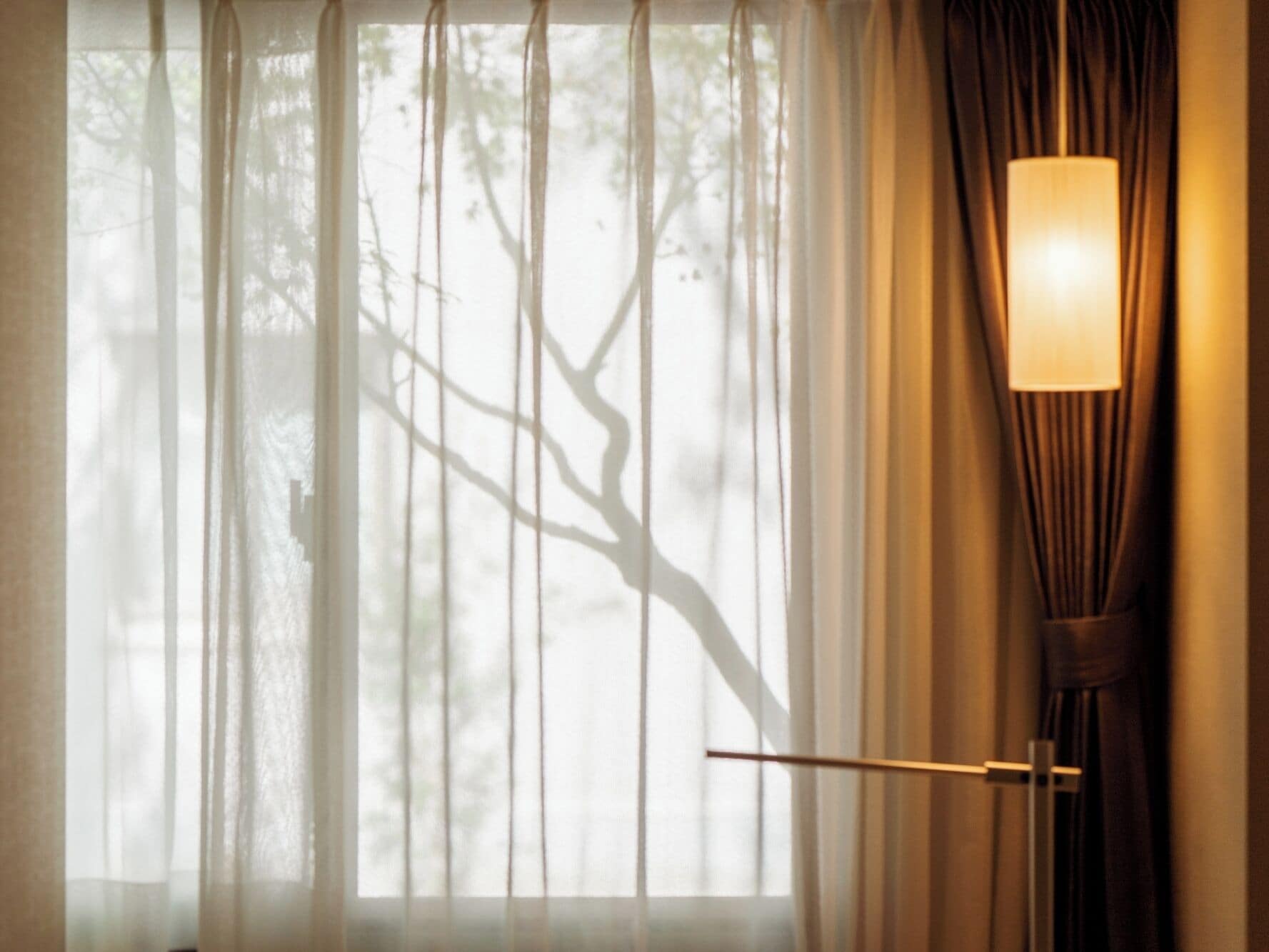 A room with a large window covered by sheer curtains, a floor lamp, and a door with a horizontal handle.