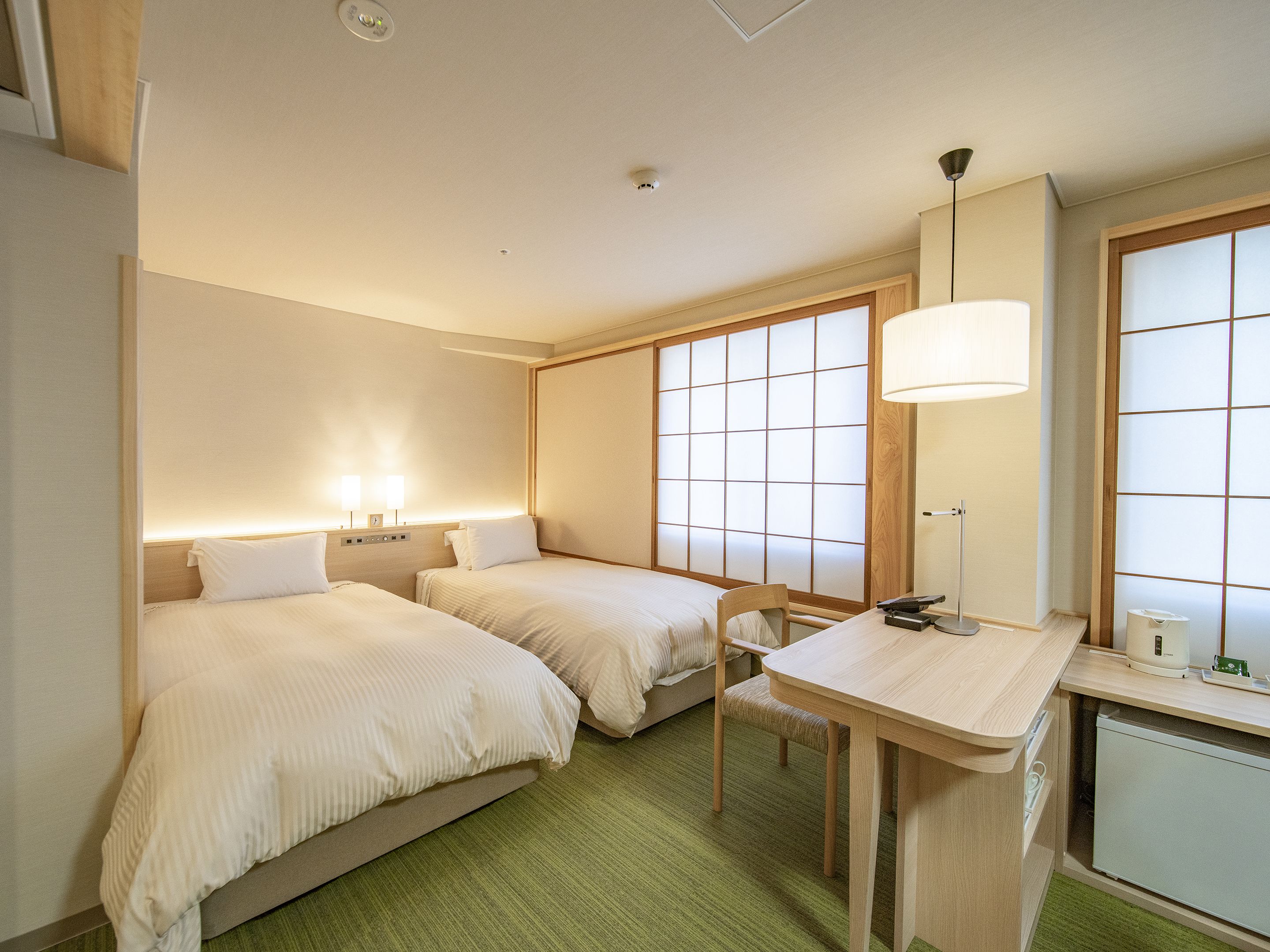 Een hotelkamer met twee bedden, een bureau, een stoel, een lamp en een raam met uitzicht.