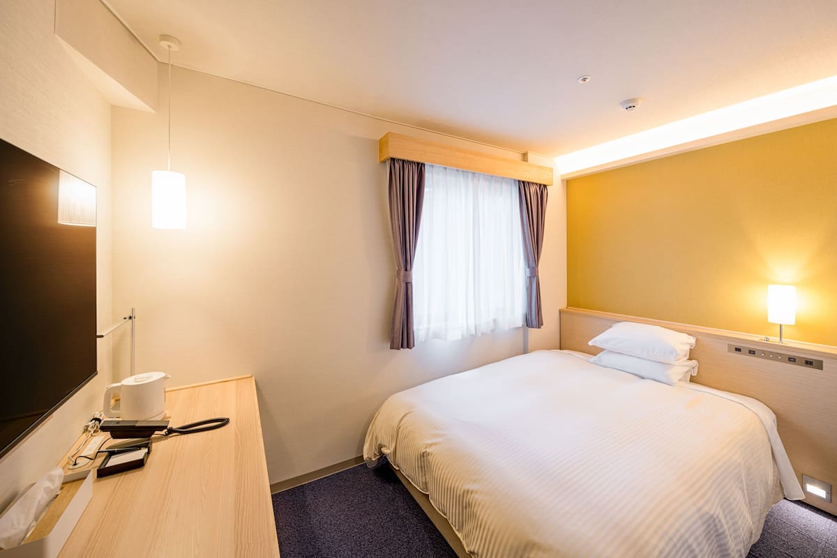 A hotel room with a bed, a desk with a telephone, a lamp, and a window with curtains.