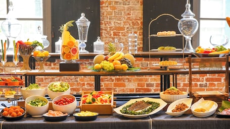 A buffet with various food items and drinks, including fruits, salads, and seafood, set against a brick wall backdrop.