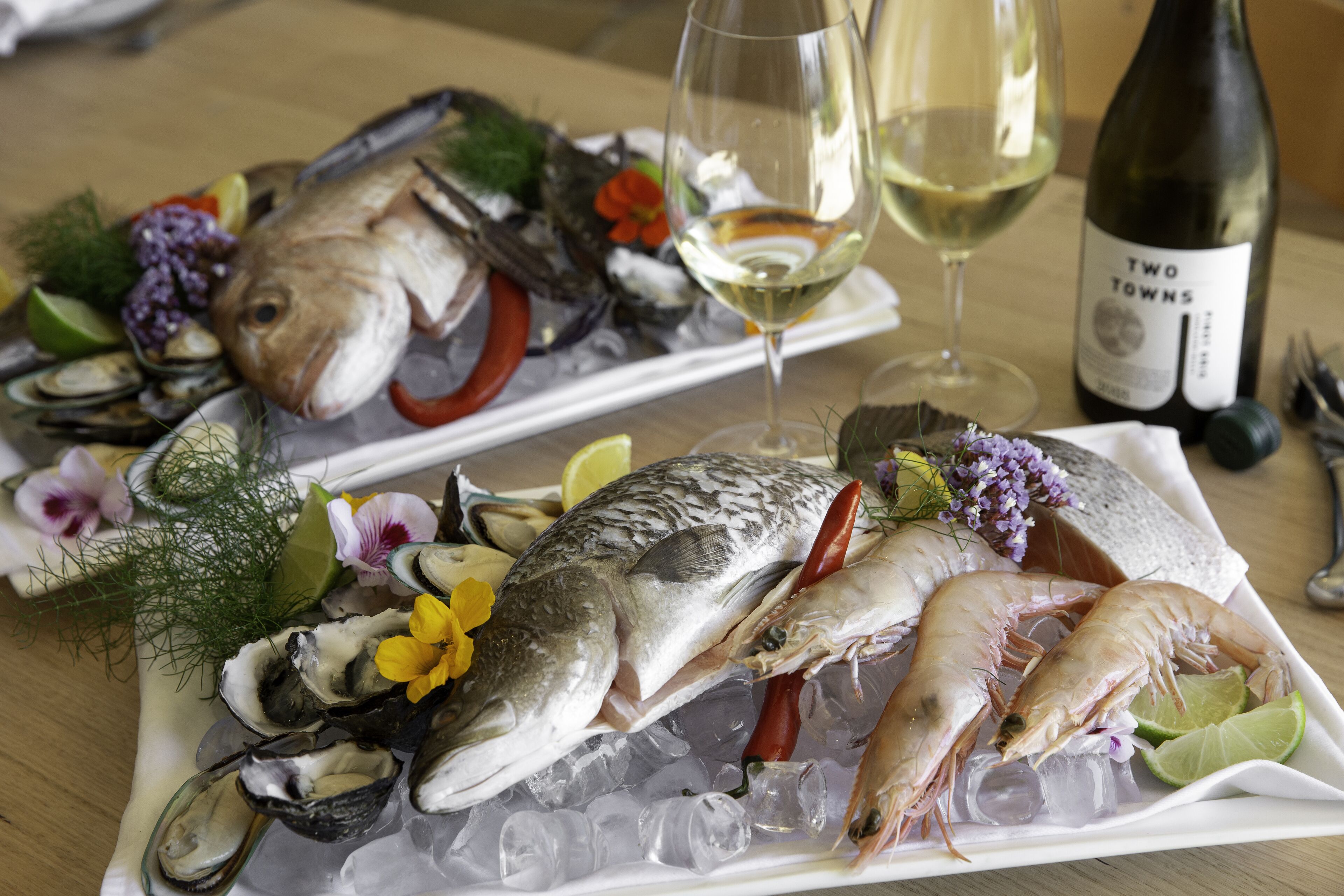 A seafood platter with oysters, shrimp, and fish on ice, accompanied by wine glasses and a bottle of wine.