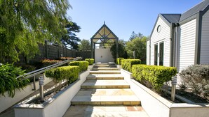 A pathway leading to a building with a triangular roof structure, surrounded by well-maintained hedges and a variety of plants.